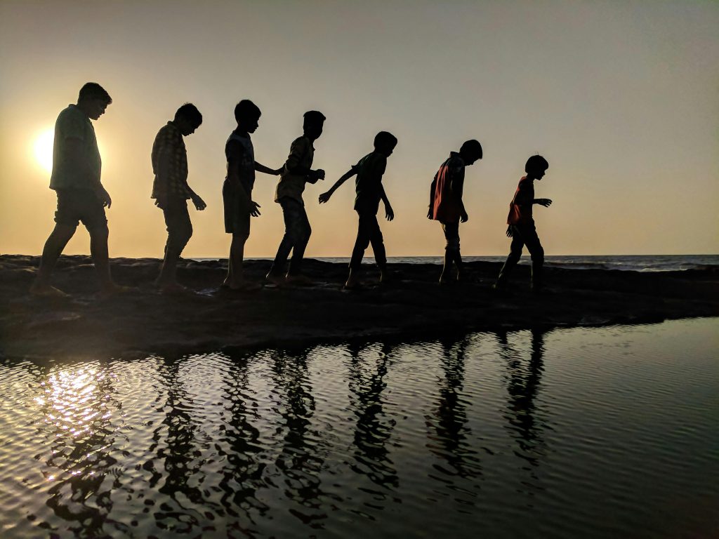 A group of children walk along the shoreline, silhouetted against a sunset, reflecting in the water.